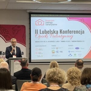 Sala konferencyjna wypełniona uczestnikami, na mównicy dr Mariusz Sałamacha, na tablicy interaktywnej tytuł konferencji “II Lubelska Konferencja Opieki Paliatywnej”