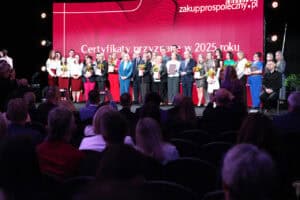 Duża grupa nagrodzonych osób, ubranych w różnorodne, formalne stroje, stoi w rzędzie na scenie. Większość z nich trzyma dyplomy i bukiety kwiatów. Za nimi znajduje się duży, czerwony ekran z białym napisem: „Certyfikaty przyznane w 2025 roku” oraz adresem strony zakupprospołeczny.pl. Na pierwszym planie widoczne są pierwsze rzędy publiczności siedzącej w ciemnej sali konferencyjnej