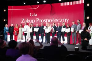 Duża grupa, około dwudziestu nagrodzonych osób stoi na scenie, trzymając certyfikaty lub dyplomy. Za nimi znajduje się ogromny, czerwony ekran z białym napisem „Gala Zakup Prospołeczny 2025”. Uczestnicy ubrani są w różnorodne, eleganckie stroje. Na pierwszym planie widoczna jest publiczność, oglądająca ceremonię z widowni