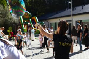 Ujęcie z bliska. Kobieta w czarnej koszulce z napisem "TRENER", odwrócona plecami, tworzy ogromną bańkę mydlaną za pomocą dwóch patyków połączonych sznurkiem. W tle, na słonecznym placu przy białym budynku, widać grupę dzieci i dorosłych oraz duży, kolorowy, dmuchany zamek