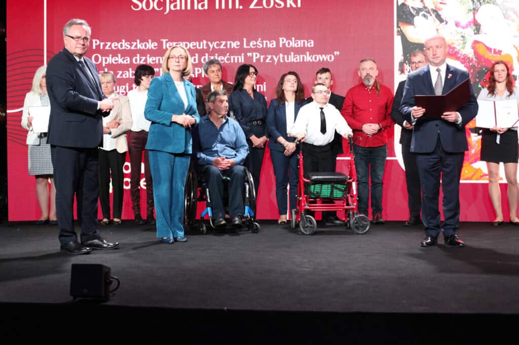 Duża grupa nagrodzonych osób stoi na scenie podczas gali. W grupie znajduje się mężczyzna na czarnym wózku inwalidzkim oraz inny mężczyzna trzymający przed sobą chodzik z siedziskiem. Na pierwszym planie po lewej stoi mężczyzna w granatowym garniturze (Zbigniew Wojciechowski), a po prawej inny mężczyzna w garniturze trzyma dużą, rozłożoną teczkę (Piotr Breś). W tle widoczny jest duży, czerwony ekran z częściowo widocznym napisem „Przedszkole Terapeutyczne Leśna Polana”