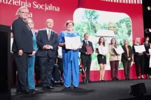 Grupa nagrodzonych osób stoi na scenie podczas gali. Kobieta w centrum, ubrana w jasnoniebieski kombinezon, trzyma duży dyplom. Obok niej stoją gospodarze wydarzenia (Zbigniew Wojciechowski, Małgorzata Romanko oraz Piotr Breś). Pozostałe osoby również trzymają certyfikaty. W tle widoczny jest duży, czerwony ekran z częściowo widocznym napisem „Spółdzielnia Socjalna” oraz dużym zdjęciem zielonego terenu z drewnianym budynkiem