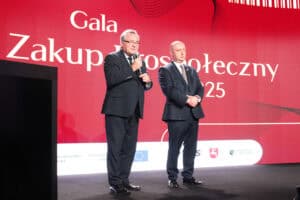 Dwóch mężczyzn w garniturach stoi na scenie. Mężczyzna po lewej (Zbigniew Wojciechowski - Radny Sejmiku Województwa Lubelskiego), w okularach, przemawia do mikrofonu, trzymając go w prawej ręce. Mężczyzna po prawej (Piotr Breś - Wicemarszałek Województwa Lubelskiego od 2024) stoi obok niego ze skrzyżowanymi rękami. W tle widoczny jest duży, czerwony ekran z białym napisem: „Gala Zakup Prospołeczny 2025” oraz logo różnych instytucji w dolnej części