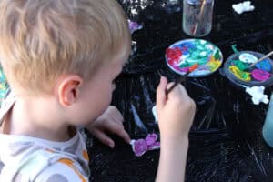 Ujęcie z góry. Chłopiec trzyma pędzelek do malowania, pochyla się nad kolorową figurką. W tle widać pojemniki z kleksami kolorowych farb i fragment słoika z pędzelkami. To wszystko znajduje się na czarnej, foliowej nawierzchni