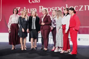 Grupa uśmiechniętych kobiet – pracowniczek ROPS w Lublinie – ubranych w eleganckie, różnorodne stroje, stoi razem na scenie podczas gali. Ich stroje obejmują m.in. sukienki w kropki, kostiumy w kolorze bordowym i czerwonym. W tle widoczny jest duży, czerwony ekran z częściowo widocznym napisem "Gala Zakup Prospołeczny"
