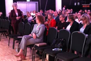 Sala konferencyjna. Na pierwszym planie siedzi kobieta w szarym garniturze, zwrócona do przodu. Za nią, w kolejnych rzędach, siedzi publiczność. Kobieta w czerwonej marynarce, trzymająca mikrofon, jest w trakcie przemówienia. Kobieta w bordowym garniturze (Marta Rak) stoi po lewej stronie, obserwując publiczność. W tle widoczny jest zegar cyfrowy, pokazujący 00:01:00