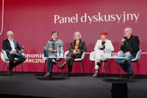Pięć osób siedzi na białych fotelach na scenie, biorąc udział w panelu dyskusyjnym. Wszyscy trzymają mikrofony. Za panelistami znajduje się duży, czerwony ekran z napisem „Panel dyskusyjny”