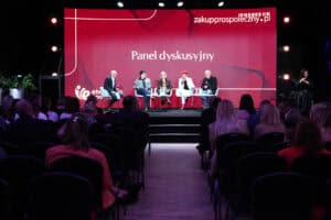 Szerokie ujęcie sali konferencyjnej. Na scenie pięć osób siedzi w białych fotelach. Za nimi znajduje się duży, czerwony ekran z białym napisem „Panel dyskusyjny” i adresem „zakupprospołeczny.pl”. Po prawej stronie sceny siedzi tłumacz języka migowego. Na pierwszym planie widoczne są rzędy krzeseł z publicznością siedzącą tyłem do obiektywu