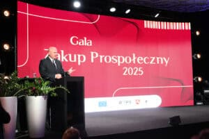 Mężczyzna (Jarosław Stawiarski) w czarnym garniturze i białej koszuli stoi za mównicą na scenie, przemawiając do widowni. Za nim znajduje się duży, czerwony ekran z białym napisem „Gala Zakup Prospołeczny 2025”. W dolnej części ekranu widoczne są logotypy, w tym flagi Unii Europejskiej. Po lewej stronie sceny stoją duże, srebrne donice z zielonymi roślinami