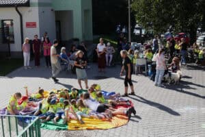 Słoneczne, szerokie ujęcie plenerowej imprezy na brukowanym placu przy budynku. Na pierwszym planie, w centrum, grupa dzieci skupionych w okręgu, w jasnych, odblaskowych kamizelkach leży na dużej, okrągłej, tęczowej chuście animacyjnej. Za matą, na środku placu, stoi kilka osób dorosłych. Po lewej stronie, pod ścianą budynku i po prawej, stoją grupy dorosłych