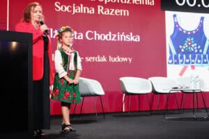 Na scenie stoi mała dziewczynka w tradycyjnym, polskim stroju ludowym (wianek z kwiatów na głowie, zielona spódniczka w czerwone róże, haftowany gorset). Obok niej stoi kobieta w czerwonej marynarce, trzymająca mikrofon. W tle widoczny jest duży, czerwony ekran z częściowo widocznym napisem „Spółdzielnia Socjalna Razem”, „Elżbieta Chodzińska” i informacją „serdak ludowy”. Po prawej stronie ekranu wyświetlone jest zbliżenie haftowanego gorsetu