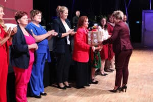 Grupa osób stoi na drewnianej podłodze w rzędzie. Kobieta w bordowym kostiumie (Marta Rak - Zastępca Dyrektora ROPS w Lublinie) stojąca z prawej strony podaje dłoń kobiecie w jaskrawoczerwonej marynarce, która trzyma duży, zawinięty prezent przewiązany wstążką. Inne kobiety, ubrane w kolorowe stroje (niebieskie, różowe, granatowe), stoją w rzędzie, oklaskując lub obserwując ten moment. W tle widoczne jest ciemne oświetlenie sceniczne.