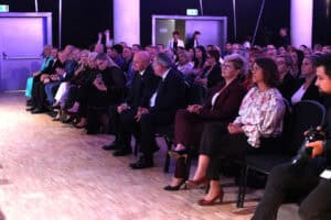 Rząd gości, ubranych w eleganckie stroje, siedzi na czarnych krzesłach podczas wydarzenia. W pierwszym rzędzie widoczne są dwie kobiety: jedna w bordowym garniturze (Marta Rak), a druga w jasnej, kwiecistej bluzce. Mężczyźni w garniturach siedzą obok nich. W tle za siedzącymi widoczne są drzwi i stojący ludzie