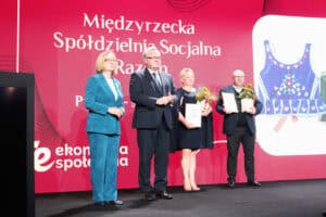 Cztery osoby – dwie kobiety i dwóch mężczyzn – stoją na scenie przed dużym, czerwonym ekranem z napisem „Międzyrzecka Spółdzielnia Socjalna Razem”. Kobieta po lewej (Małgorzata Romanko - Dyrektor ROPS) ma na sobie turkusowy kostium, stoi w towarzystwie mężczyzny w garniturze (Zbigniew Wojciechowski). Kobieta w środku i mężczyzna po prawej trzymają dyplomy i bukiety kwiatów. Na ekranie, po prawej stronie, wyświetlony jest duży obraz haftowanej kamizelki