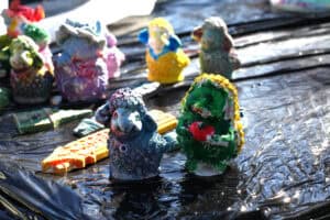 Na czarnym tle stoją kolorowe figurki zwierząt, malowane ręcznie. Na pierwszy plan wysuwają się dwie figurki: niebiesko-szarej owcy i jeża w kolorach: zielonym, żółtym i czerwonym