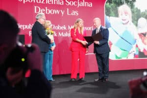 Cztery osoby na scenie podczas ceremonii wręczenia nagród. Mężczyzna w garniturze (Piotr Breś) ściska dłoń kobiety w jaskrawoczerwonym kostiumie biznesowym, jednocześnie wręczając jej duży, rozłożony dyplom. Obok nich, po lewej, stoi inna para: mężczyzna w garniturze (Zbigniew Wojciechowski), trzymający kwiaty i kobieta w turkusowym kostiumie (Małgorzata Romanko). W tle widoczny jest duży, czerwony ekran z napisem „Spółdzielnia Socjalna Dębowy Las” oraz ramka z uśmiechniętym dzieckiem