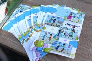 Widok z góry na ulotki rozłożone w formie wachlarza na powierzchni drewnopodobnej. Na ulotkach są kolorowe obrazki, typu historyjki. Po prawej wyróżnia się napis: „Tacy sami”