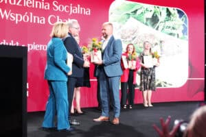 Ceremonia wręczenia nagród na scenie. Mężczyzna w granatowym garniturze (Zbigniew Wojciechowski) stoi przed mężczyzną w jasnoszarym garniturze, który trzyma dyplom i bukiet kwiatów. Po lewej stronie stoi kobieta w turkusowym kostiumie (Małgorzata Romanko). W tle widać dwie inne nagrodzone kobiety, również trzymające kwiaty i dyplomy. Za nimi znajduje się duży, czerwony ekran z napisem „Spółdzielnia Socjalna Wspólna Chata” i zdjęciem produktu