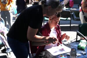 Przy stoliku siedzi chłopiec w koszuli w kratkę, a nad nim pochyla się kobieta, która dłońmi otacza dłoń młodego człowieka. Trzyma on pędzel, nad leżącą na białym tle figurką pomalowaną częściowo na różowo