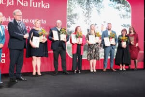 Grupa ośmiu dorosłych osób stoi w rzędzie na scenie, pozując po odebraniu nagród. Wszyscy z wyjątkiem jednego mężczyzny w garniturze po lewej (Zbigniew Wojciechowski) trzymają dyplomy i bukiety słoneczników. W tle widoczny jest duży, czerwono-biały ekran z napisem „NaTurka” oraz duży obraz drzew i przyrody po prawej stronie