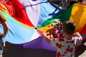 Zbliżenie na zabawę z wielobarwną chustą animacyjną wypełnioną kolorowymi piłkami. Stojący tyłem mały chłopiec w szarej wzorzystej koszulce trzyma brzeg chusty. Piłki podskakują, a wokół widać części sylwetek innych dzieci i dorosłych. Zabawa prowadzona jest na brukowanej nawierzchni w słoneczny dzień