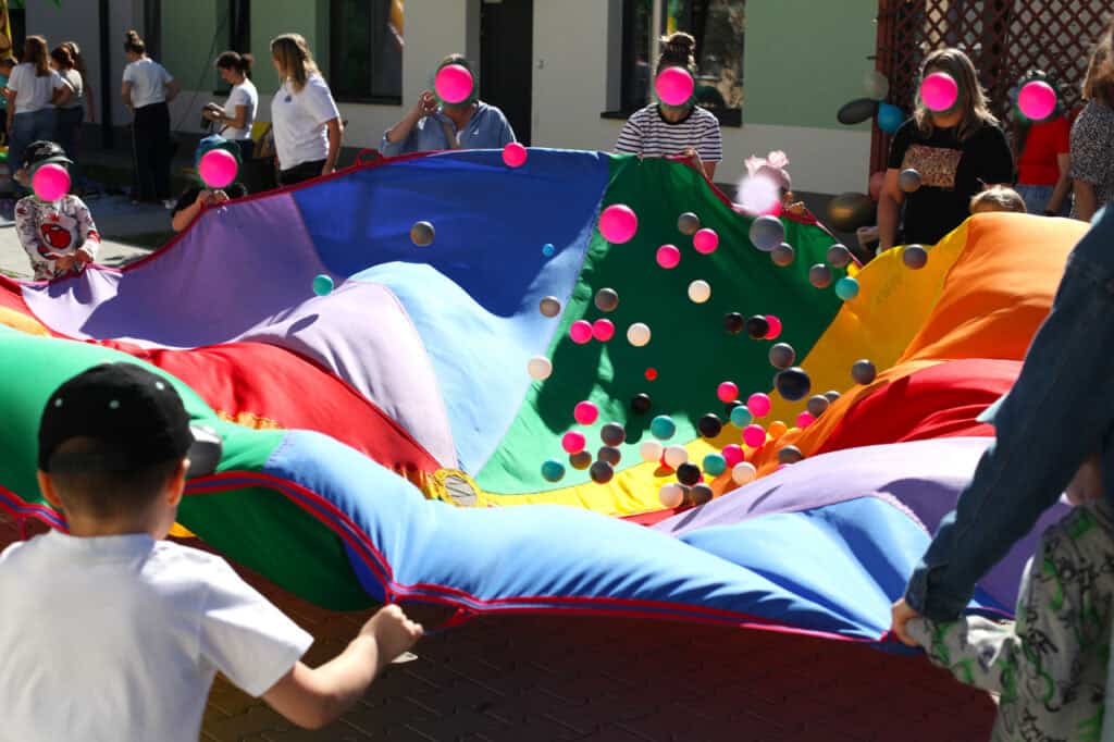 Zabawa wielobarwną chustą animacyjną. Za brzegi materiału trzymają dzieci i dorośli, tworząc koło. Na powierzchni materiału podskakują kolorowe piłeczki (różowe, białe, szare, czarne). Chusta składa się z kolorowych segmentów: żółty, zielony, pomarańczowy, niebieski, czerwony). W tle ściana budynku