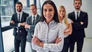 Kobieta w białej koszuli stoi ze skrzyżowanymi ramionami przed trzema kolegami i jedną koleżanką w strojach biznesowych, którzy pozują za nią. Ich twarze są niewyraźne.