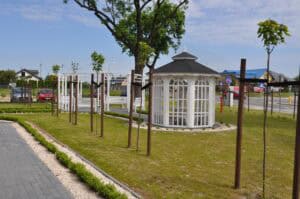 Na pierwszym planie biała, ażurowa altana stojąca pośrodku starannie przyciętego trawnika, na którym w równych odstępach posadzono młode drzewa liściaste. Tuż za altaną znajdują się cztery białe ławki ustawione w kręgu. W tle widoczne są zaparkowane samochody, zabudowa uliczna i elementy infrastruktury drogowej