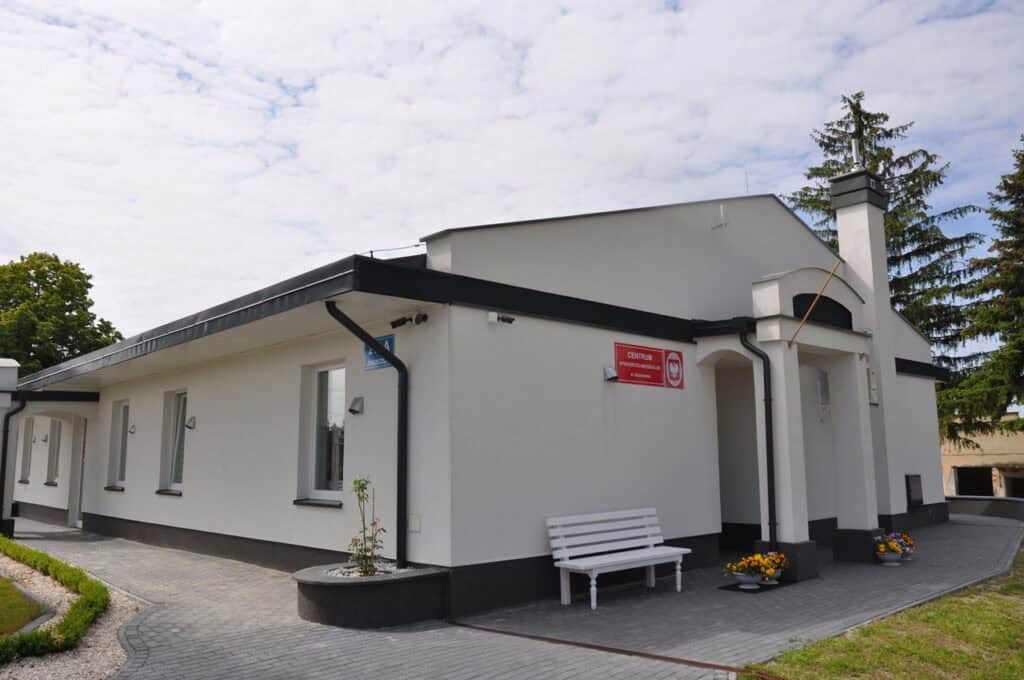 Parterowy, biały budynek z czerwoną tabliczką ‘Centrum Opiekuńczo-Mieszkalne w Jaszczowie’. Przed kolumnowym wejściem stoją doniczki z kwiatami i biała ławeczka. Otoczenie zadbane – kostka brukowa, trawnik i drzewa w tle