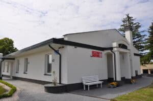 Parterowy, biały budynek z czerwoną tabliczką ‘Centrum Opiekuńczo-Mieszkalne w Jaszczowie’. Przed kolumnowym wejściem stoją doniczki z kwiatami i biała ławeczka. Otoczenie zadbane – kostka brukowa, trawnik i drzewa w tle