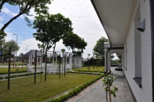 Zagospodarowany teren przed parterowym, białym budynkiem. Przy samej elewacji zadaszony pas szarej kostki, dalej wąski pas białego grysu z niskimi krzaczkami bukszpanu. Przestrzeń otacza starannie przycięty trawnik, na którym w równych odstępach posadzono młode drzewa liściaste. W centralnej części znajdują się cztery białe ławki ustawione w kręgu, a tuż obok biała, ażurowa altana