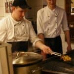 Pomieszczenie kuchenne. Dwóch młodych mężczyzn w bluzach kucharskich i czapkach z daszkiem stoi przy ruszcie. Pierwszy z lewej trzyma szczypce i obraca mięso. Na pierwszym planie, (nieostry) duży stalowy gar z przykrywką