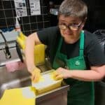 Pomieszczenie kuchenne. Młoda kobieta z bardzo krótko ostrzyżonymi włosami, ubrana w czarny t-shirt i zielony fartuch z plakietką imienną "OLA", stoi przy stalowym zlewozmywaku. Sięga rękami do pojemnika z żółtymi ściereczkami