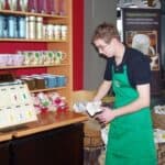 Wnętrze kawiarni Starbucks. Po lewej fragment regału z kubkami. Obok stoi lekko pochylony mężczyzna w czarnej koszulce polo i zielonym fartuchu, trzyma w rękach jasny przedmiot – prawdopodobnie ścierkę lub papier. W tle kosze i torby jutowe w stojakach, roll-up ze zdjęciem kubka kawy oraz po prawej przeszklona witryna z ladą i wysokimi krzesłami barowymi