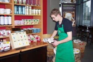 Wnętrze kawiarni Starbucks. Po lewej fragment regału z kubkami. Obok stoi lekko pochylony mężczyzna w czarnej koszulce polo i zielonym fartuchu, trzyma w rękach jasny przedmiot – prawdopodobnie ścierkę lub papier. W tle kosze i torby jutowe w stojakach, roll-up ze zdjęciem kubka kawy oraz po prawej przeszklona witryna z ladą i wysokimi krzesłami barowymi
