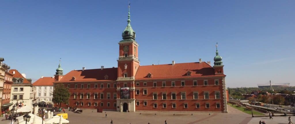 Widok zewnętrzny Zamku Królewskiego w Warszawie, z charakterystyczną czerwoną ceglaną fasadą, dachem krytym dachówką i wieżą zegarową.