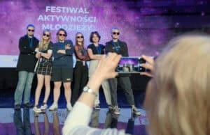 Grupa młodych osób stojąca na scenie w okularach przeciwsłonecznych. Ktoś na pierwszym planie robi zdjęcie telefonem.