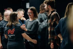 Grupa ludzi rozmawia i wchodzi w interakcję w towarzystwie. Jedna z kobiet ma na sobie koszulkę z nadrukiem „Nieprzetarty szlak” na plecach.