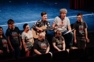 Grupa osób na scenie z ciemnoniebieskim tłem. Część z nich nosi koszulki z napisem 'Akademia Artystyczna'. Kobieta z brązowymi włosami gra na gitarze i śpiewa. Obok niej stoi starszy mężczyzna z siwymi, kręconymi włosami, trzymający papiery.