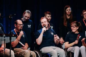 Chór stoi na scenie przed ciemną kurtyną. Większość nosi identyczne, granatowe koszulki z białym motywem anielskich skrzydeł. Niektórzy członkowie chóru są na wózkach inwalidzkich. Dwie osoby trzymają mikrofony, śpiewając. Obok nich kobieta trzyma mały, czerwony instrument z dzwoneczkiem, potrząsając nim podczas występu.