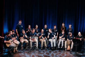 Grupa 17 osób, w tym dwie osoby na wózkach, występująca na scenie na tle ciemnoniebieskiej kurtyny, grają na różnych instrumentach, mają na sobie pasujące do siebie ciemne koszulki z emblematem, niektórzy śpiewają do mikrofonów, a zakonnica gra na ukulele.