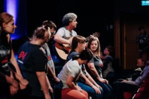 Mężczyzna gra na gitarze akustycznej przed siedzącą publicznością.