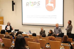 Sala wykładowa. Kobieta (Anna Bieganowska-Skóra) w ciemnym żakiecie, stoi po lewej stronie, trzyma w rękach kartkę i przemawia do widowni. Po prawej, przy ekranie projekcyjnym, znajdują się dwie kobiety i dwóch mężczyzn. W tle, na dużym, jasnym ekranie widać logo ROPS (Regionalny Ośrodek Polityki Społecznej) Lublin, a także informację: „Zadanie publiczne jest finansowane ze środków PFRON przyznanych przez Samorząd Województwa Lubelskiego”. Większość kadru zajmują tyły głów i plecy słuchaczy siedzących w rzędach na jasnych, drewnianych krzesłach.