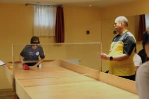 Dwie osoby na hali sportowej podczas gry w showdown. W centrum, kobieta w ciemnym sportowym stroju i opasce na oczy (zasłaniającej widoczność) trzyma rakietkę i jest pochylona nad drewnianym stołem z bandami i przezroczystą, środkową przegrodą. Po prawej, mężczyzna w żółto-czarnej koszulce, z opaską na szyi i zegarkiem na nadgarstku, stoi, obserwując grę