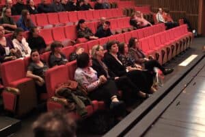 Długa perspektywa rzędów czerwonych foteli na widowni teatru, z wieloma osobami siedzącymi i czekającymi na rozpoczęcie wydarzenia. W pierwszym planie widoczna grupa kobiet (m.in. Anna Bieganowska-Skóra, Marzena Rząsowska, Joanna Olszewska)