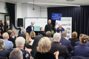 Sala konferencyjna. Na pierwszym planie widać tył głów i plecy publiczności, która siedzi i bije brawo. Na scenie stoją cztery osoby: z lewej mężczyzna w muszce, w centrum mężczyzna w czarnej koszuli trzymający niebieską tabliczkę z napisem „KROK ZA KROKIEM ZAMOŚĆ", obok niego siedzi na wózku mężczyzna, a po prawej kobieta przy mikrofonie. Z tyłu widoczne są: duży ekran z prezentacją oraz baner Stowarzyszenia Pomocy Dzieciom Niepełnosprawnym „Krok za Krokiem".