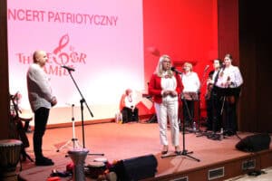 Koncert patriotyczny, scena oświetloną na czerwono. Kobieta (Dorota Jędrych-Przerwa) w czerwonym swetrze i białych spodniach przemawia na scenie do mikrofonu. Po lewej stronie stoi mężczyzna (Marek Fedor), a w tle widać zespół muzyczny i instrumenty perkusyjne leżące na podłodze