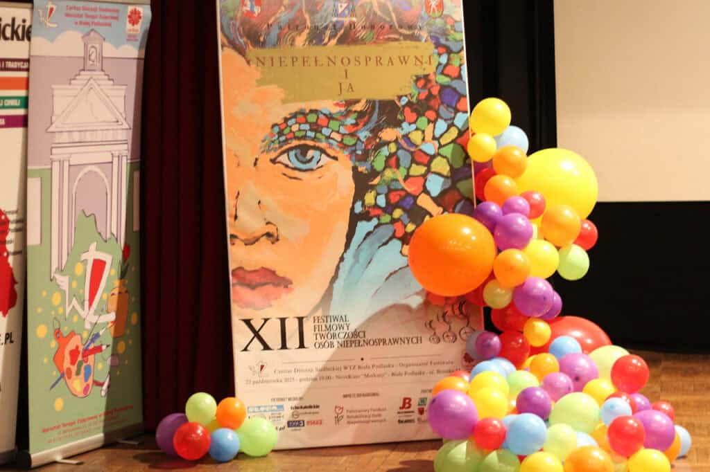 Duży plakat reklamowy XII Festiwalu Filmowego Twórczości Osób Niepełnosprawnych "Niepełnosprawni i Ja", przedstawiający wyrazisty, graficzny, kolorowy portret kobiety. Plakat otoczony jest dekoracją z kolorowych, okrągłych balonów w odcieniach żółci, pomarańczy, fioletu i czerwieni, ustawionych na drewnianej podłodze sceny