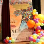 Duży plakat reklamowy XII Festiwalu Filmowego Twórczości Osób Niepełnosprawnych "Niepełnosprawni i Ja", przedstawiający wyrazisty, graficzny, kolorowy portret kobiety. Plakat otoczony jest dekoracją z kolorowych, okrągłych balonów w odcieniach żółci, pomarańczy, fioletu i czerwieni, ustawionych na drewnianej podłodze sceny