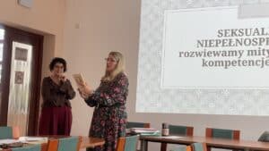 Sala wykładowa. Pomiędzy stołami stoją dwie kobiety, jedna z nich trzyma przed sobą książkę i czyta tytuł lub autora. W tle na ścianie wyświetlony jest slajd z tytułem wykładu